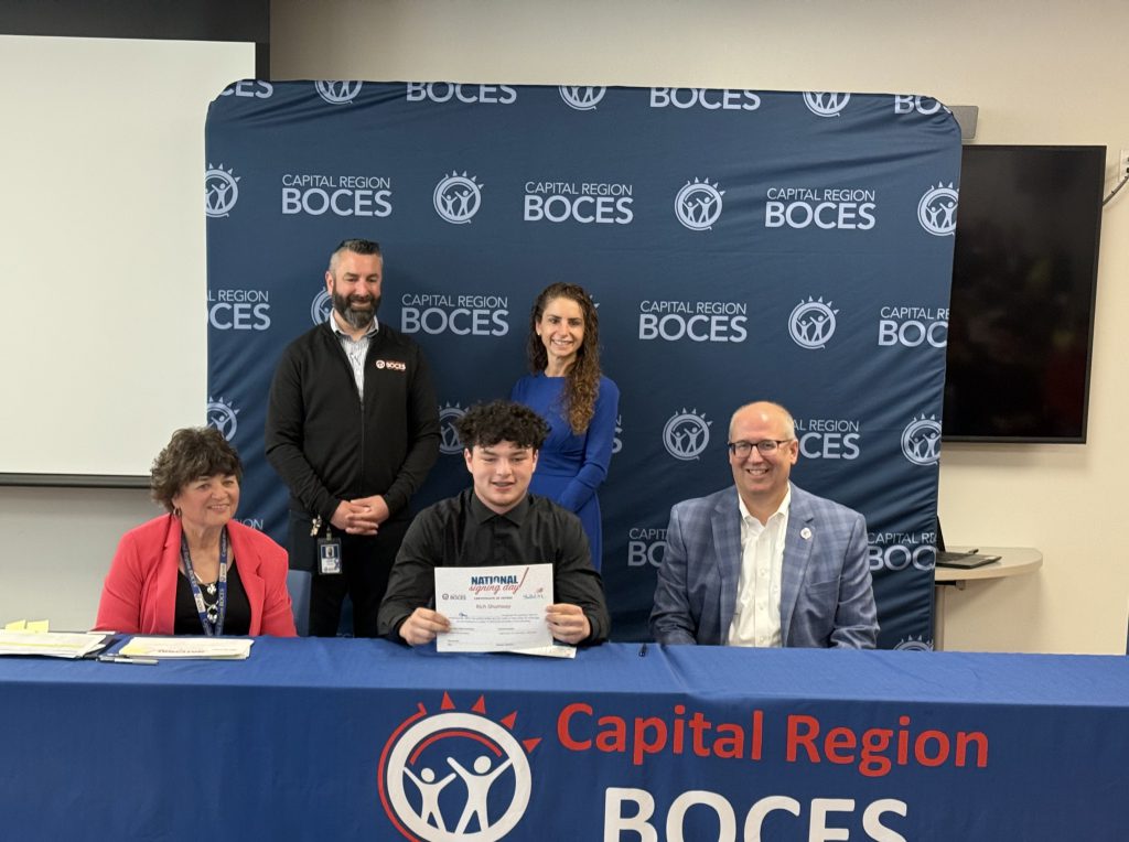 A group of five individuals is positioned around a table with a blue tablecloth that reads "Capital Region BOCES". Three individuals are seated, and two are standing behind them. The individual in the center foreground, seated, holds a certificate that says "National Signing Day"