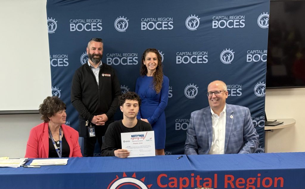 A group of five individuals is positioned around a table with a blue tablecloth that reads "Capital Region BOCES". Three individuals are seated, and two are standing behind them. The individual in the center foreground, seated, holds a certificate that says "National Signing Day"